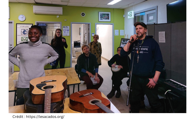 plusieurs personnes dans une pièce lors d'un atelier de musique. Un homme chante, deux autres personnes font des percussions, on voit des guitares empilées au premier plan